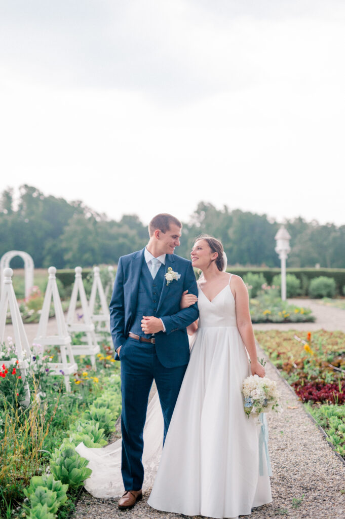 Couple smiling at each other while walking through vibrant garden beds at Smith Farm Gardens during their Connecticut summer wedding.