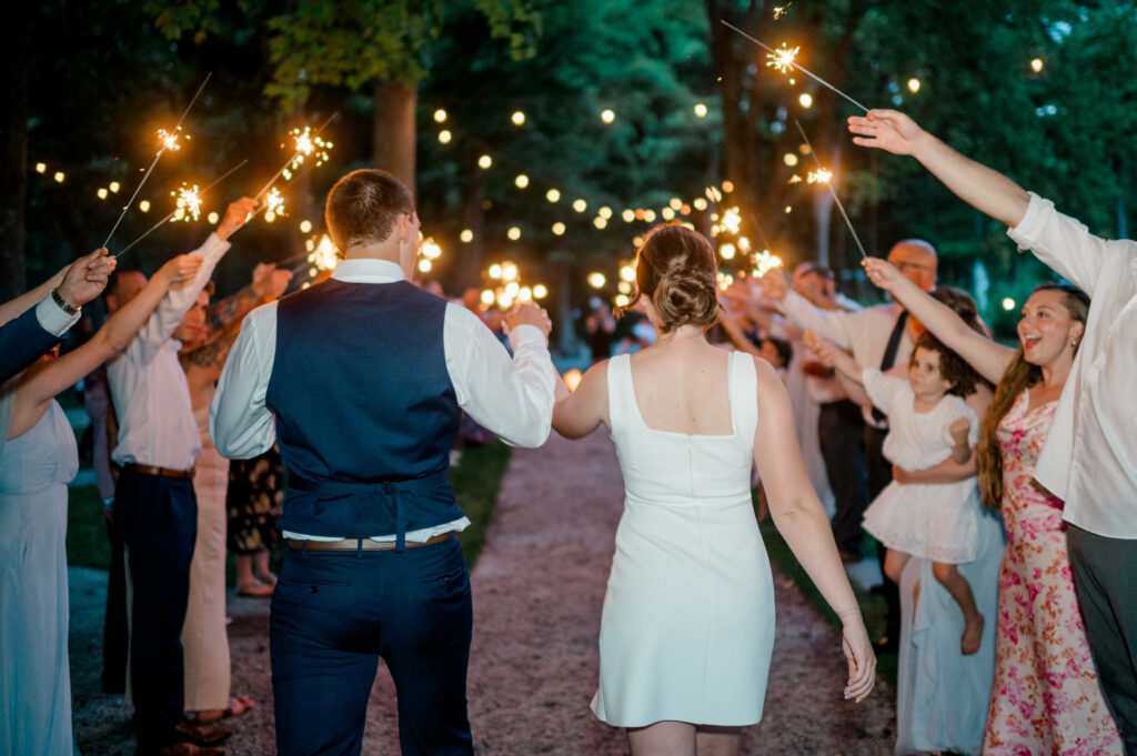 Alexis and Doug walk away hand-in-hand under a canopy of glowing sparklers, framed by cheering guests during their evening wedding sendoff.