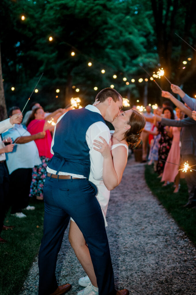 The couple shares a romantic kiss under string lights and a tunnel of sparklers, celebrating their joyful night exit at Smith Farm Gardens.