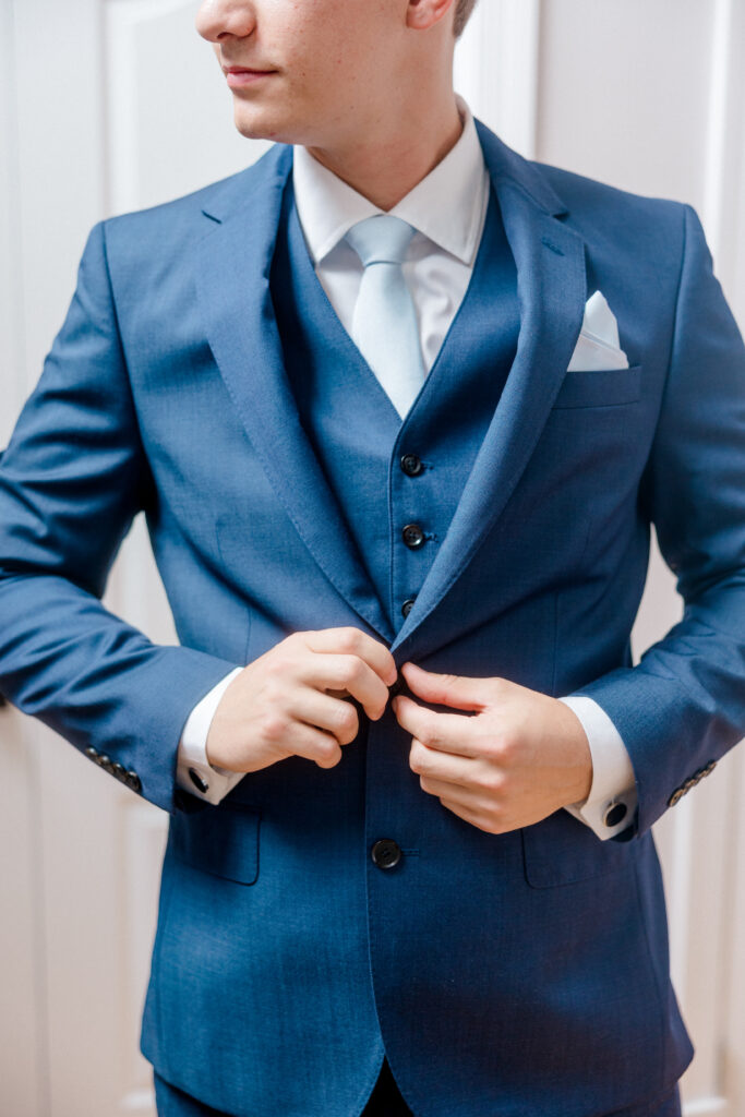Groom adjusting his navy blue suit jacket and light blue tie, readying for the ceremony with polished details.