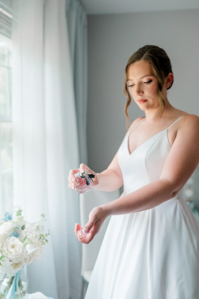 Bride spritzing perfume onto her wrist, standing beside her bouquet in the soft morning light of the getting ready suite.