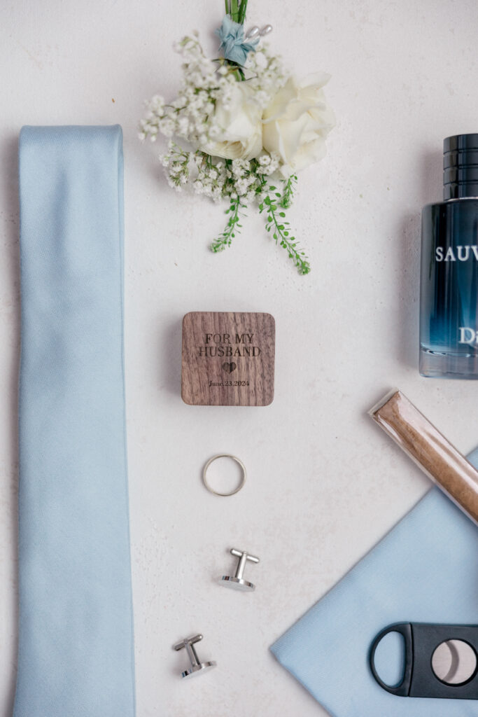 Groom’s flat lay with light blue tie, boutonnière, cufflinks, and cologne, alongside a keepsake box reading “For My Husband.”
