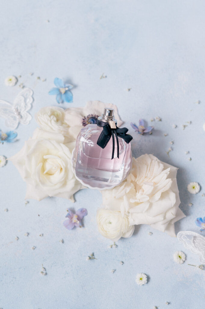 Close-up flat lay of bride’s perfume bottle surrounded by soft white roses, with delicate blue accents.