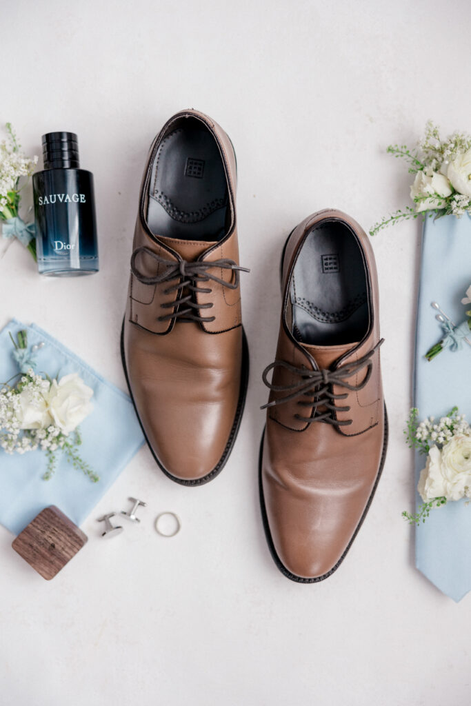 Brown dress shoes laid out with floral boutonnieres and cufflinks, styled for a clean, editorial groom detail shot.