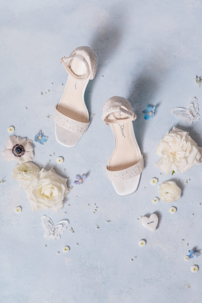 Flat lay of pearl-accented bridal heels surrounded by white garden roses and lace butterflies, styled on a soft blue background for a fine art wedding detail.