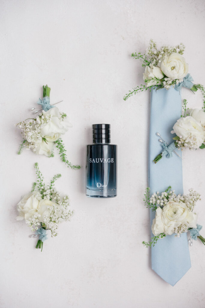 Groom’s accessories displayed with Dior Sauvage cologne, pale blue tie, and fresh boutonnieres.