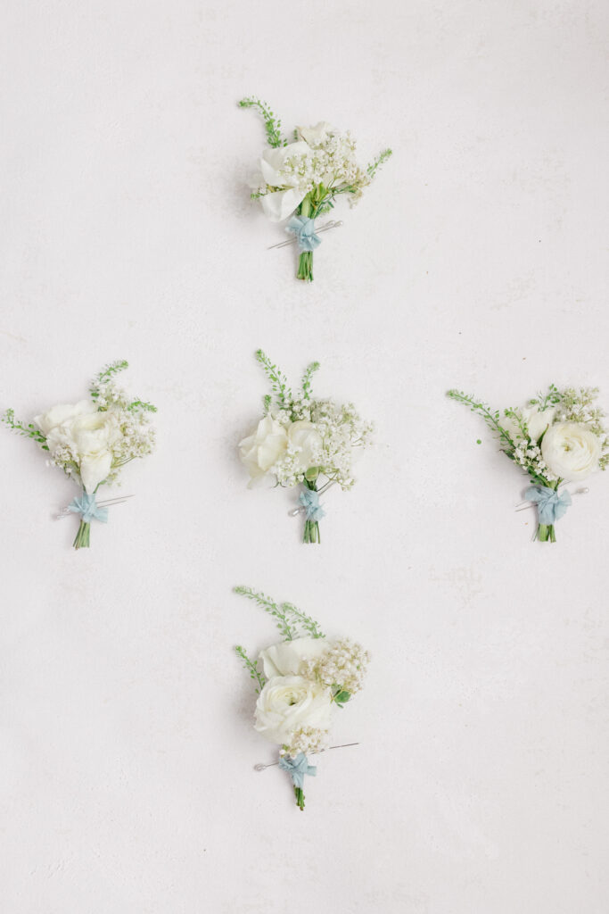 Flat lay of white boutonnieres tied with blue ribbon, styled on a clean white background.