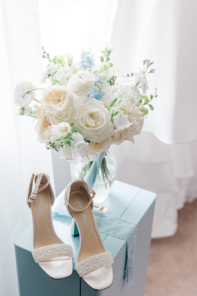 Softly lit bridal bouquet of white and pale blue florals sits beside a pair of pearl-embellished wedding heels on a powder blue pedestal.