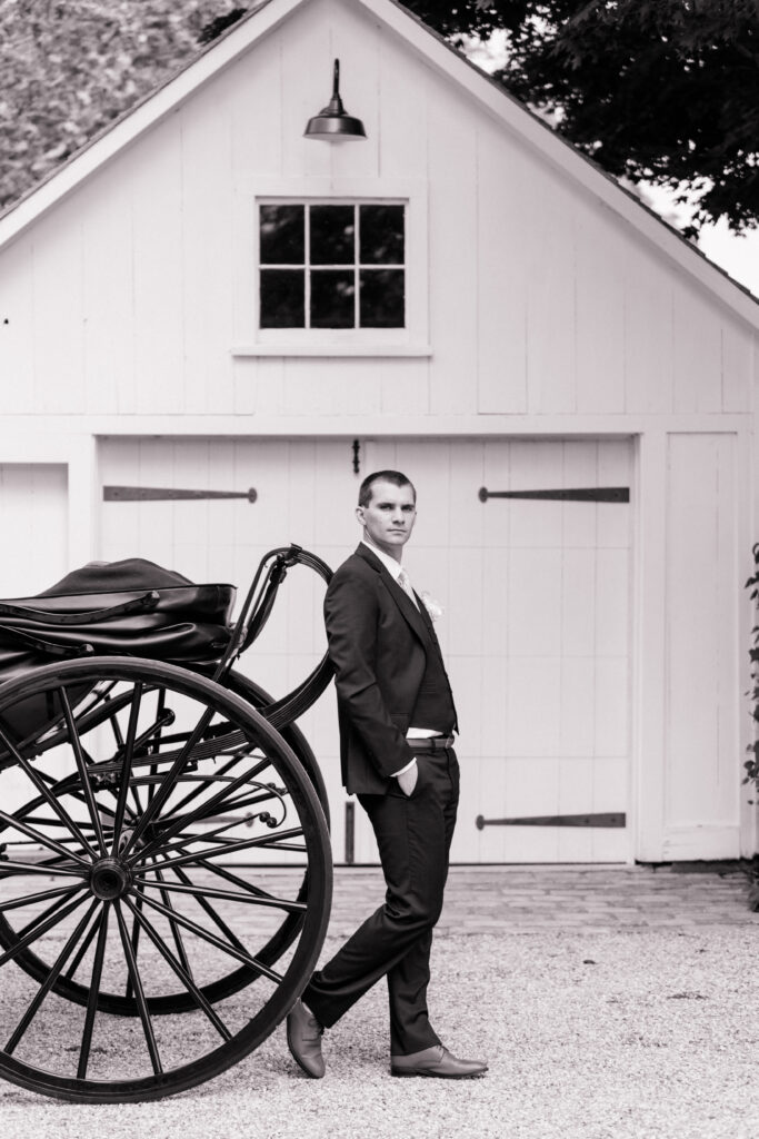 Editorial groom portrait next to vintage carriage and white barn doors at Smith Farm Gardens, CT wedding venue.