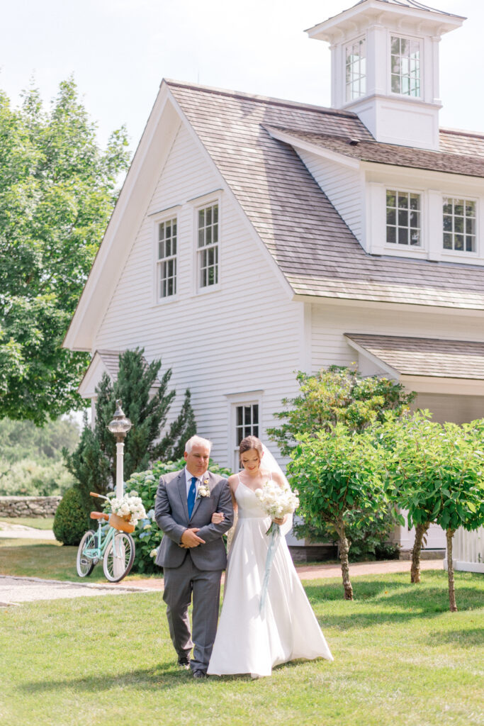Bride walks down the aisle with her father in front of the iconic white barn at Smith Farm Gardens, holding a white bouquet with soft blue ribbon.