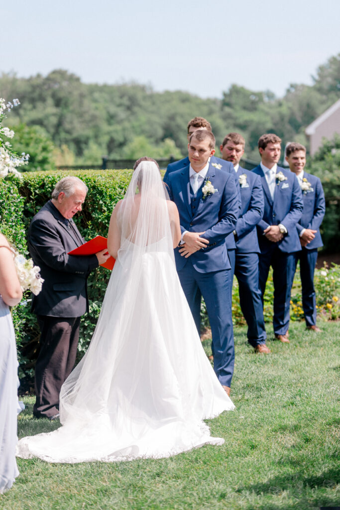 Bride and groom exchange vows under the summer sun, with the groom and groomsmen dressed in blue suits at Smith Farm Gardens.