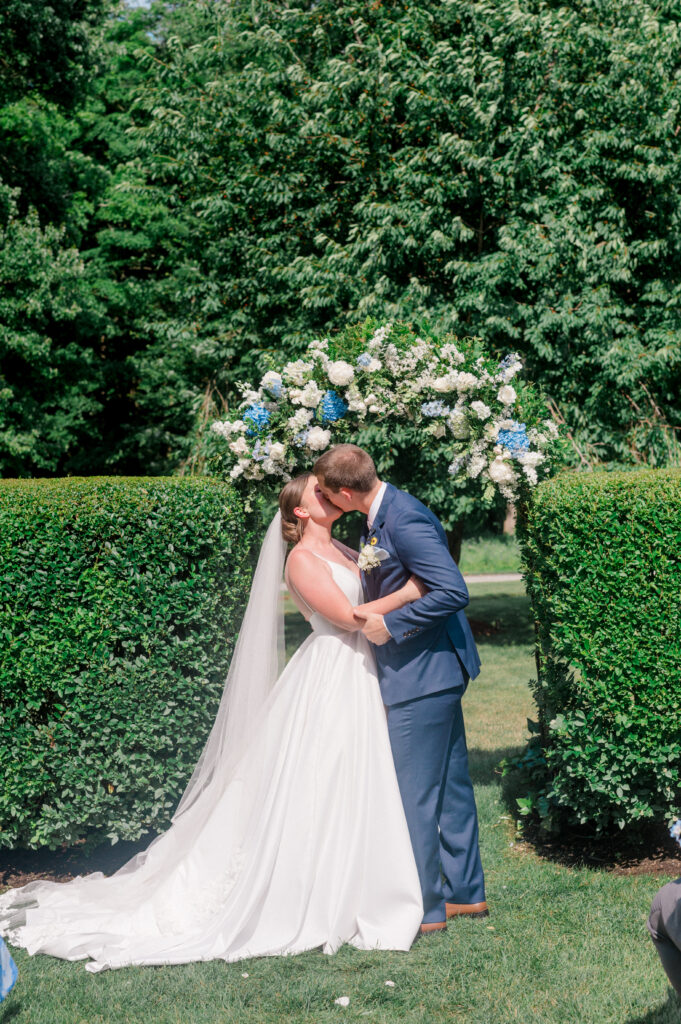 The couple shares their first kiss under a stunning blue and white flower arch at their outdoor Smith Farm Gardens ceremony.