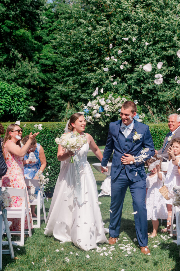 Bride and groom walk down the aisle surrounded by cheering guests and floating white flower petals in the garden at Smith Farm Gardens.