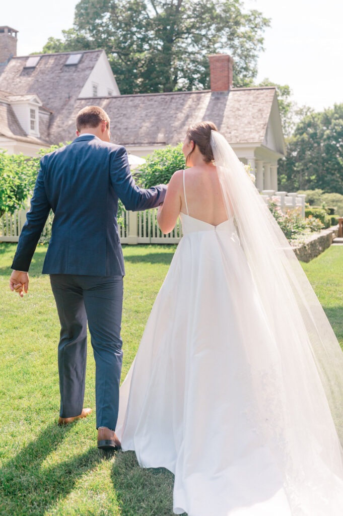 Candid moment of the bride and groom walking hand-in-hand toward their ceremony at Smith Farm Gardens, her veil trailing behind.