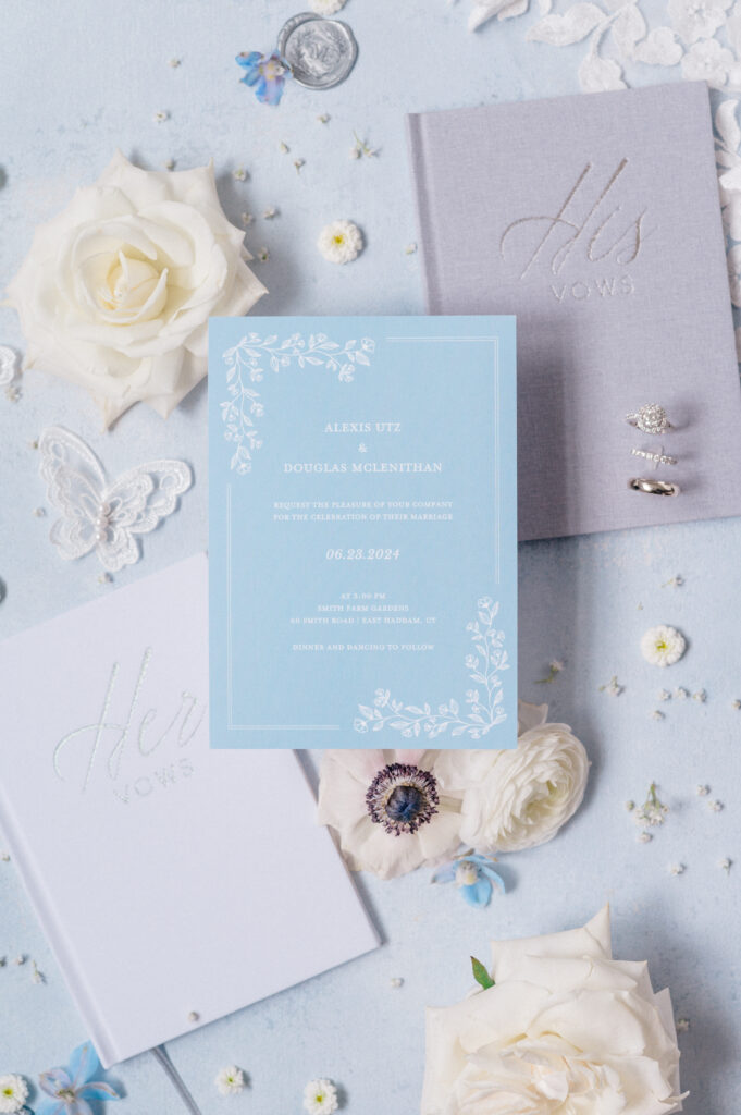 Flat lay of bride and groom’s vow books and invitation suite, styled with florals and lace butterflies on a blue linen backdrop.
