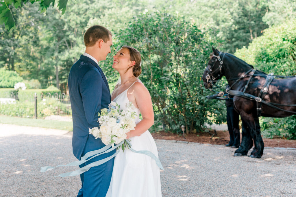 Bride and groom share a joyful gaze in front of lush greenery and a black horse-drawn carriage at Smith Farm Gardens, with the bride holding a white and blue bouquet tied with silk ribbons.