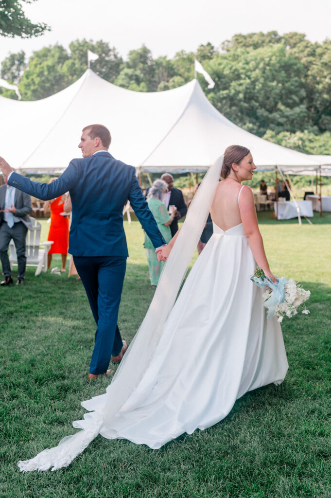 The couple strolls hand in hand across the lawn, her dress flowing behind her, framed by the sailcloth tent in the background.