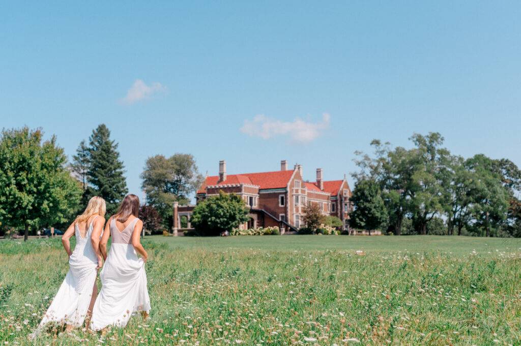 Two brides walking through a wildflower field toward Waveny House, captured in a romantic and candid moment by Jennifer Nichole & Co.
