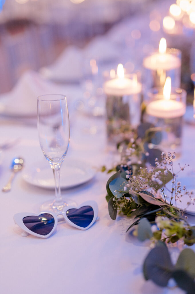 Close-up of Newport Harbor Island wedding reception table with heart-shaped sunglasses, floating candles, and white linens.