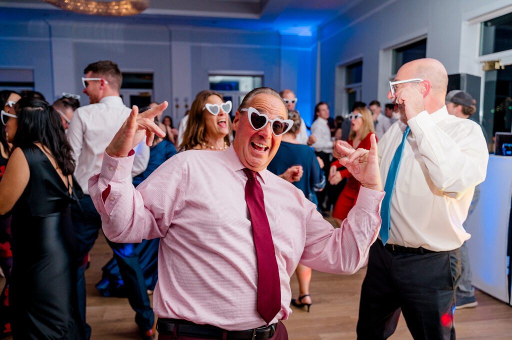 Wedding guest in pink shirt having fun in heart-shaped sunglasses at Newport Harbor Island wedding celebration.