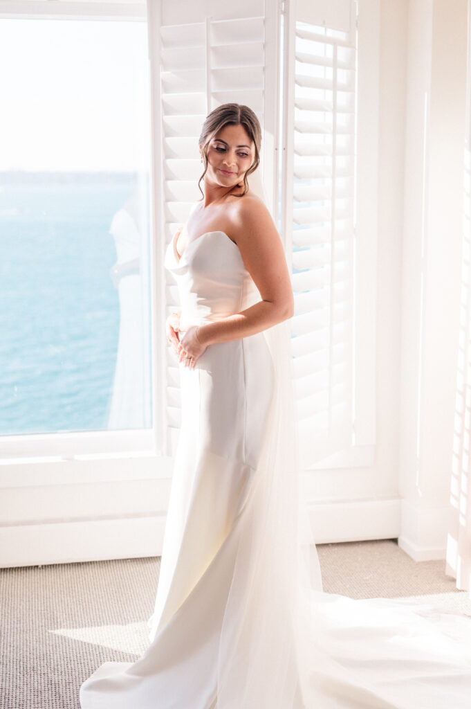 Bride Alexandra in soft light, posing by ocean-facing window before her elegant wedding at Newport Harbor Island Resort