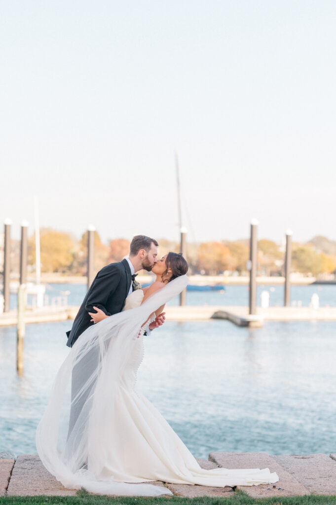 Groom dipping bride into a kiss with dramatic veil movement and marina in the background