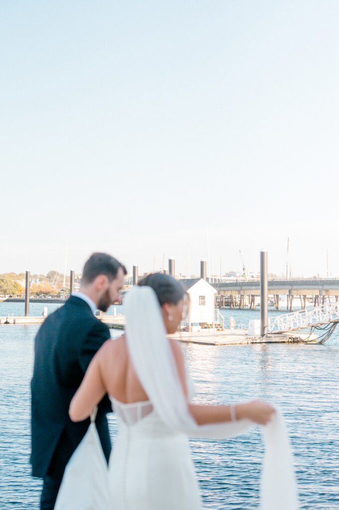 Bride and groom walk along dockside holding hands with blurred marina backdrop