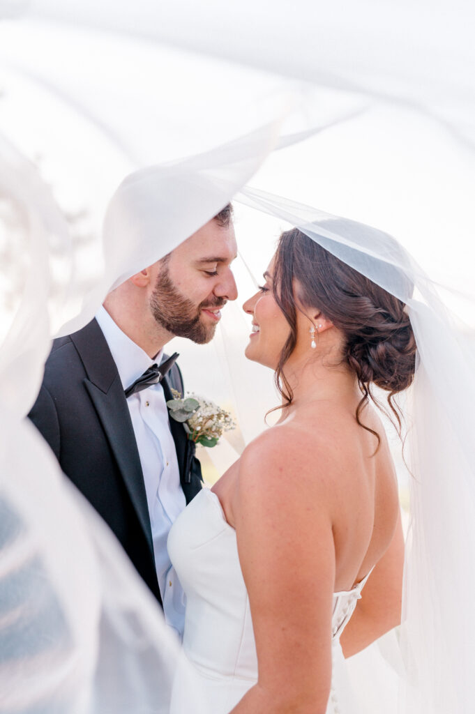 Intimate portrait under the bride’s veil as couple smiles at each other in soft evening light