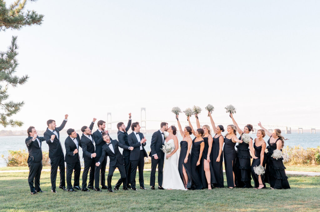 Entire wedding party in black tie cheering on newlyweds with the Newport bridge in the background.