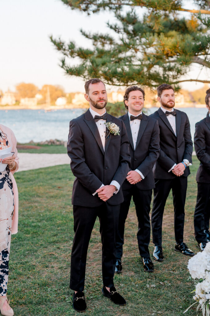 Groom stands with groomsmen in anticipation, smiling as his bride walks down the aisle at their elegant waterfront ceremony in Newport.