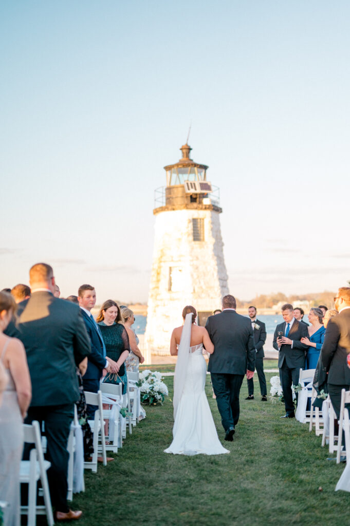 Bride walks arm-in-arm with her father down the ceremony aisle at Newport Harbor Island Resort, approaching the groom with the Goat Island lighthouse ahead.