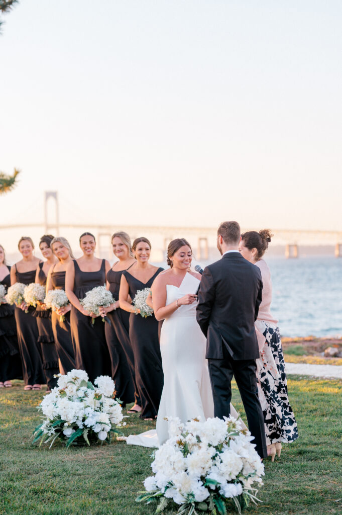 Bride reads her vows to groom while bridesmaids in black dresses holding baby’s breath bouquets stand beside her, with the iconic Newport Bridge in the background.
