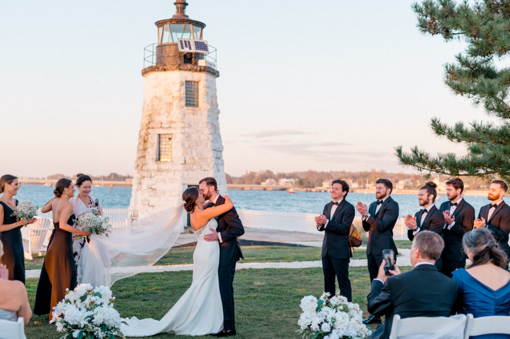 Newlyweds share their first kiss at their Newport Harbor Island Resort wedding, surrounded by cheering bridesmaids and groomsmen with ocean views and lighthouse in the background.