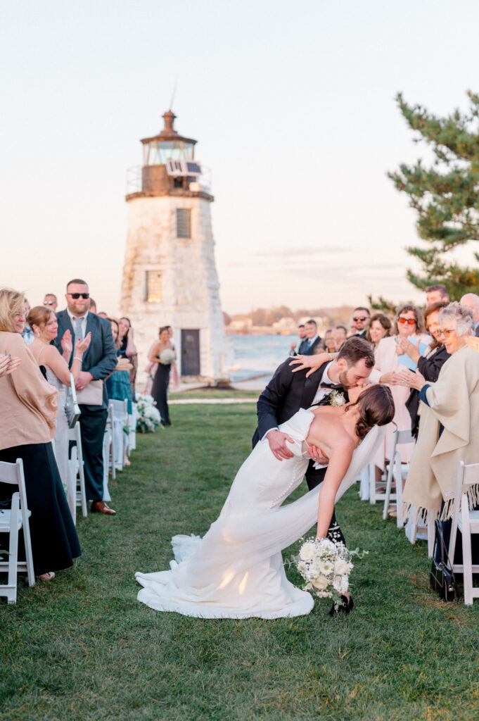 Bride and groom share a romantic kiss at the end of their waterfront wedding ceremony aisle at Newport Harbor Island Resort, with the Goat Island lighthouse behind them.