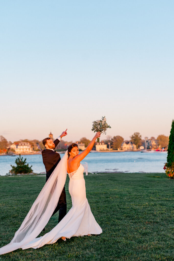 Bride and groom celebrate just after their waterfront ceremony at Newport Harbor Island Resort, both raising their arms in joy with a bouquet and a playful point toward the sky, bathed in golden hour light.