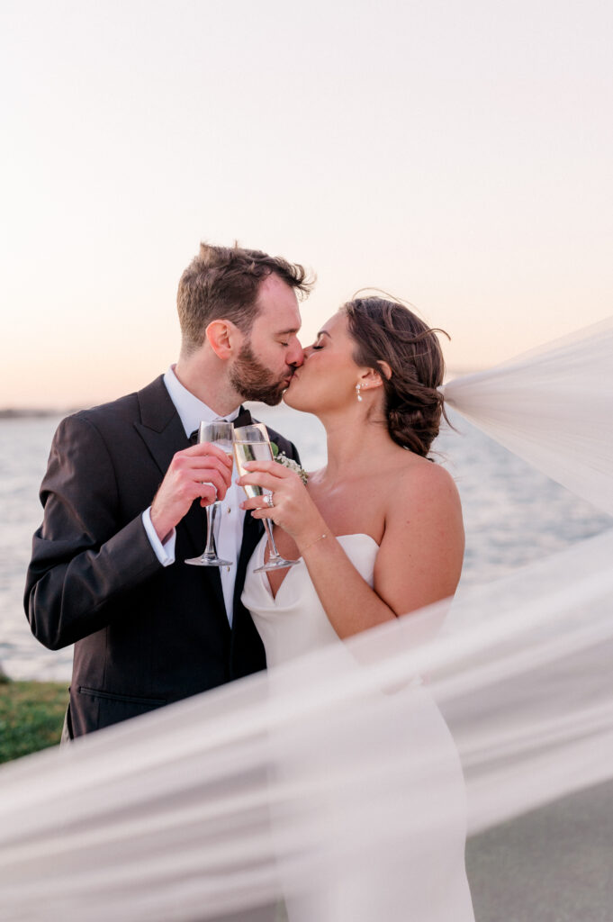 Newlyweds kiss with champagne glasses in hand and veil sweeping across frame at sunset