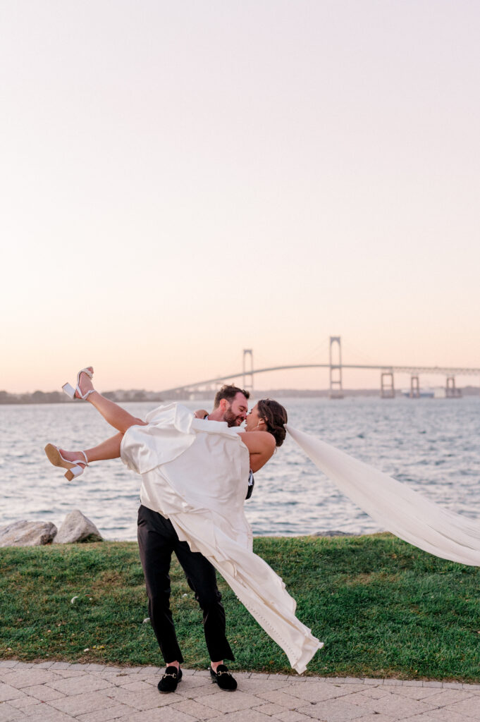 Groom lifts bride in romantic embrace near ocean with Claiborne Pell Bridge behind them