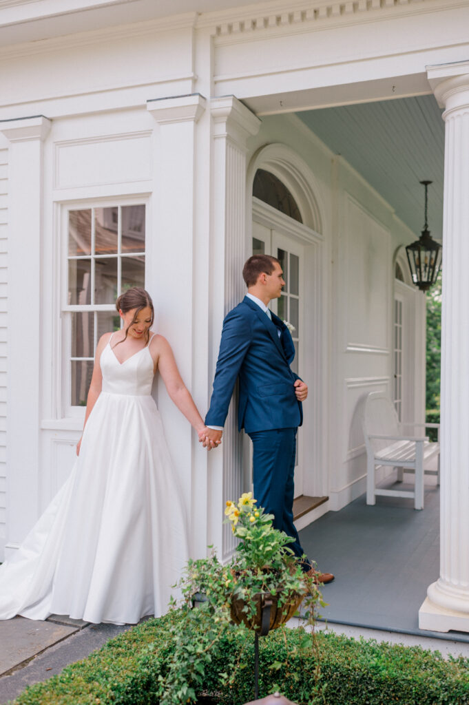 Bride and groom hold hands around white porch corner for an emotional first touch at Smith Farm Gardens wedding