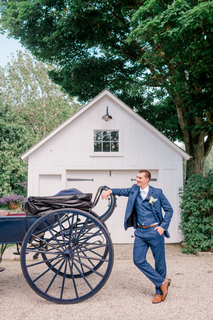 Groom in blue suit smiling and leaning casually against navy horse-drawn carriage, outside a white barn surrounded by green trees at a New England wedding.