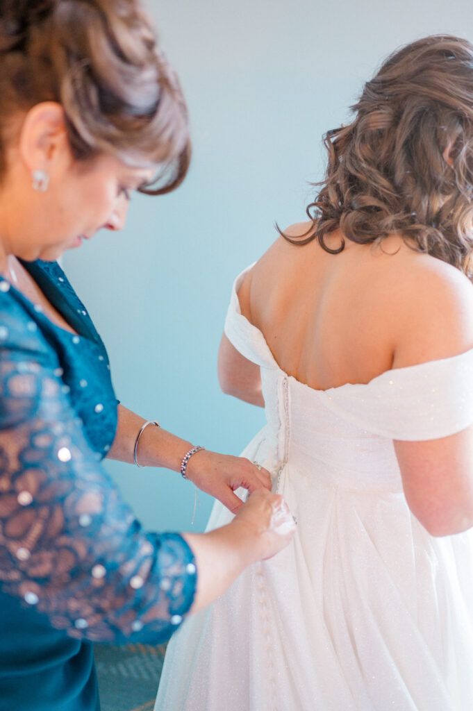 Mother helping bride button back of off-shoulder wedding gown, documenting Connecticut wedding photography timeline getting ready moments.