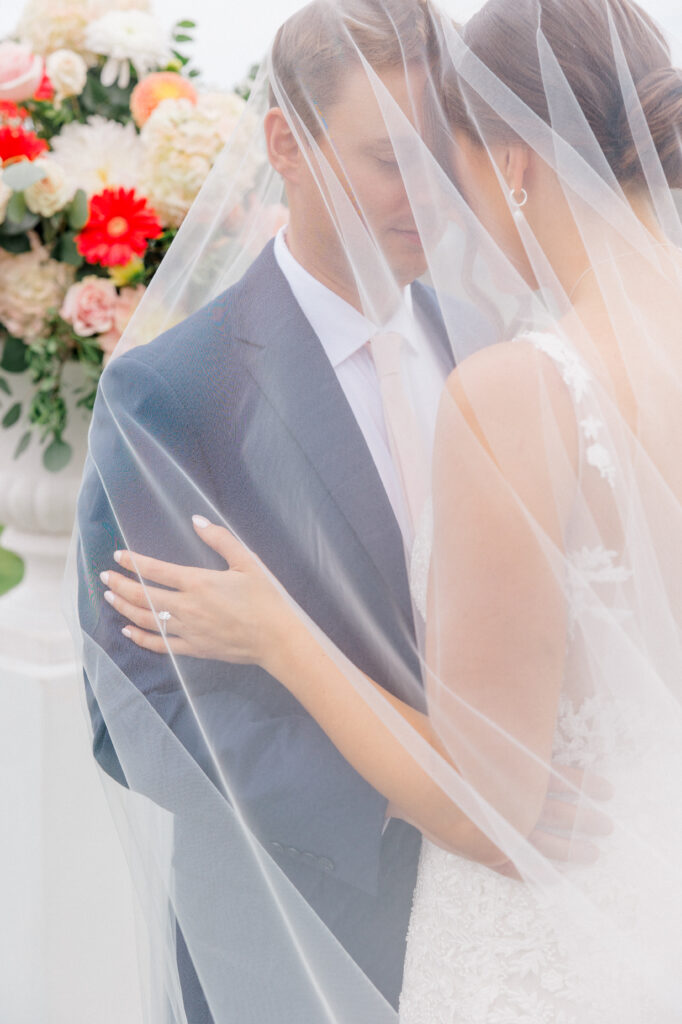 Bride and groom standing closely under bride’s veil, sharing an intimate moment, with soft focus on colorful floral backdrop at a Connecticut wedding ceremony.