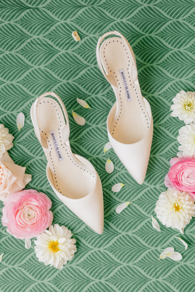 Cream-colored Manolo Blahnik wedding shoes styled on patterned green backdrop with ranunculus and dahlia petals.