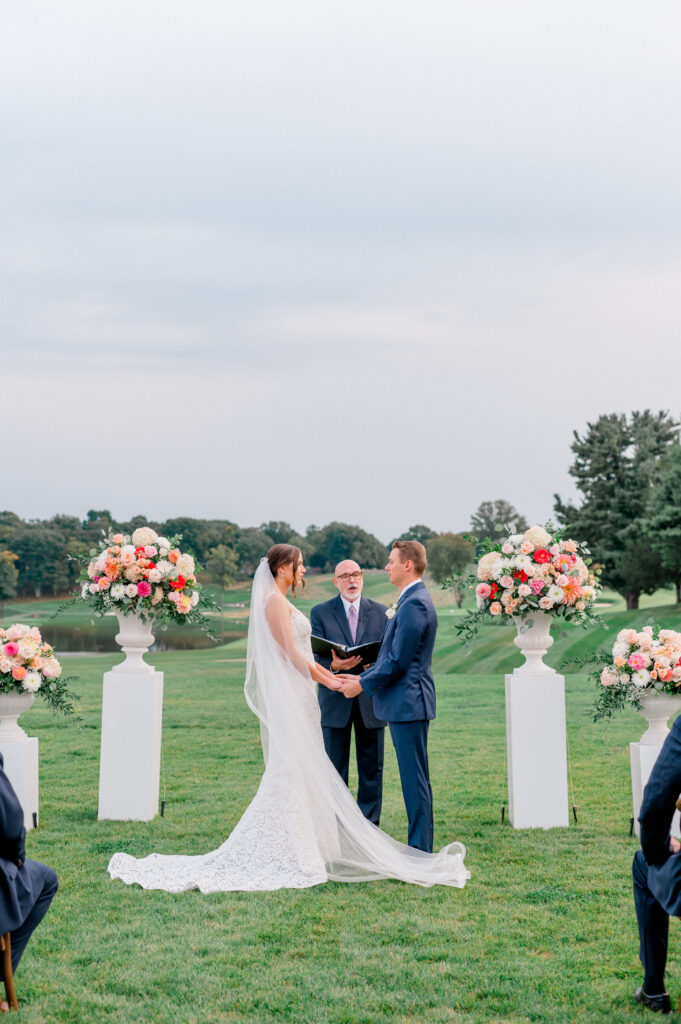 Bride and groom holding hands during outdoor Connecticut wedding ceremony, framed by vibrant floral arrangements on white pedestals during a TPC River Highlands wedding captured by Jennifer Nichole & Co.