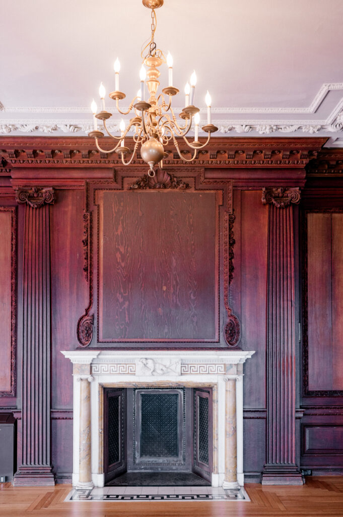 Gold chandelier above a marble fireplace surrounded by dark wood paneling at Branford House wedding ballroom