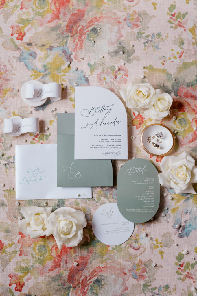Green and white wedding invitation suite styled on floral linen with vow ribbons, roses, and custom details for heirloom flatlay photography.