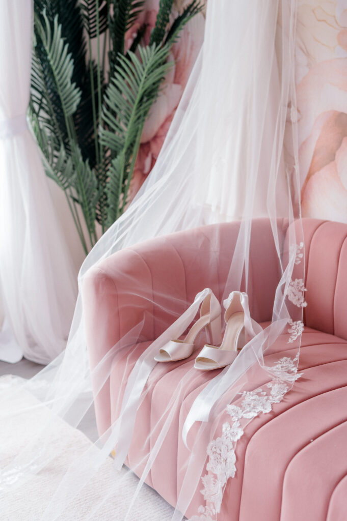 Ivory bridal heels and lace-edged veil styled over a blush velvet sofa for soft and elegant wedding morning photography details.
