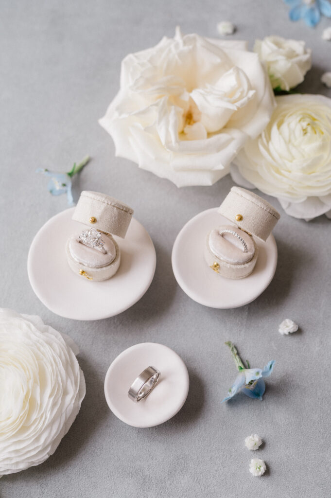 Romantic flat lay of wedding rings in velvet boxes styled with ivory roses and white ranunculus for a fine art wedding morning.