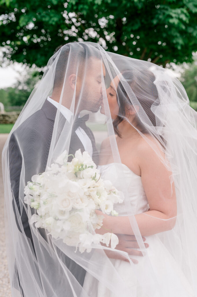 Bride and groom kiss beneath a flowing veil while holding a white floral bouquet.