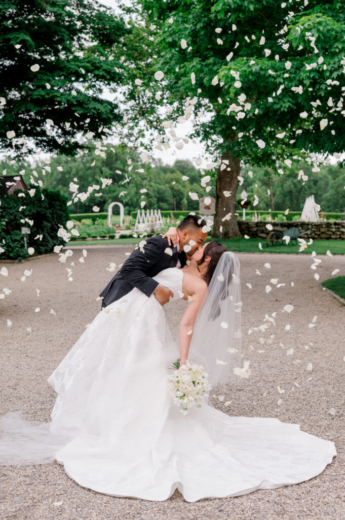Newlyweds share a romantic kiss as white flower petals fall around them at Smith Farm Gardens.