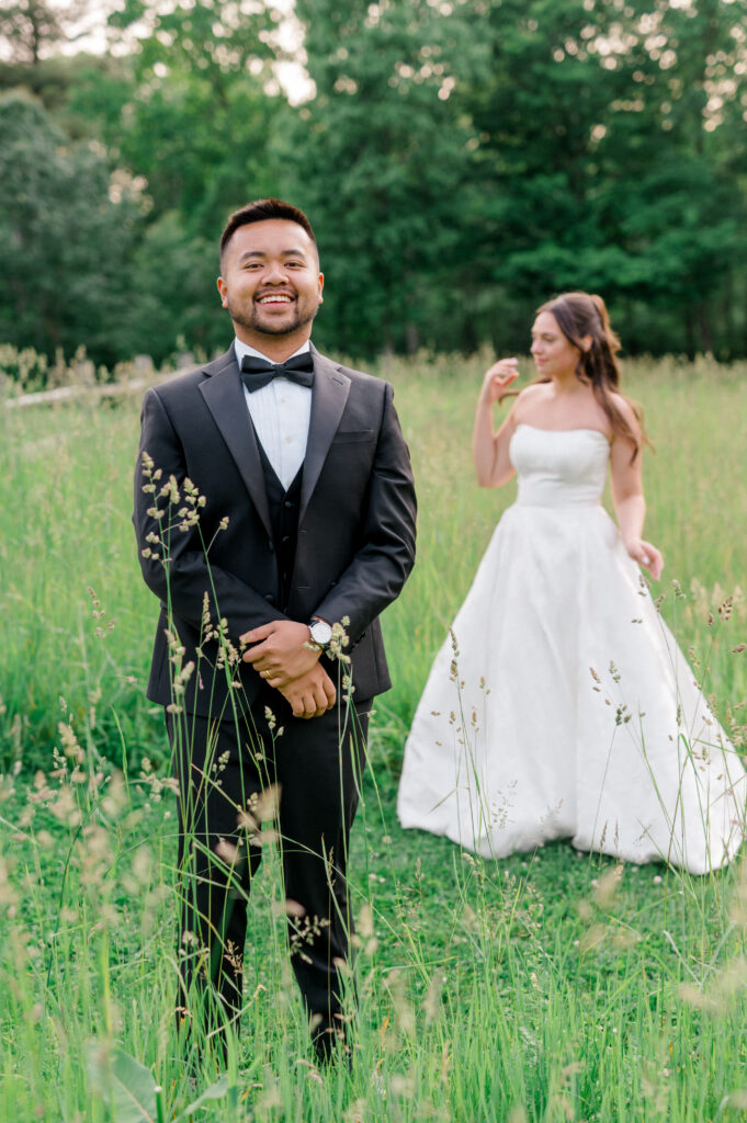 Groom smiling in a black tuxedo with his bride playfully blurred in the background of a meadow.